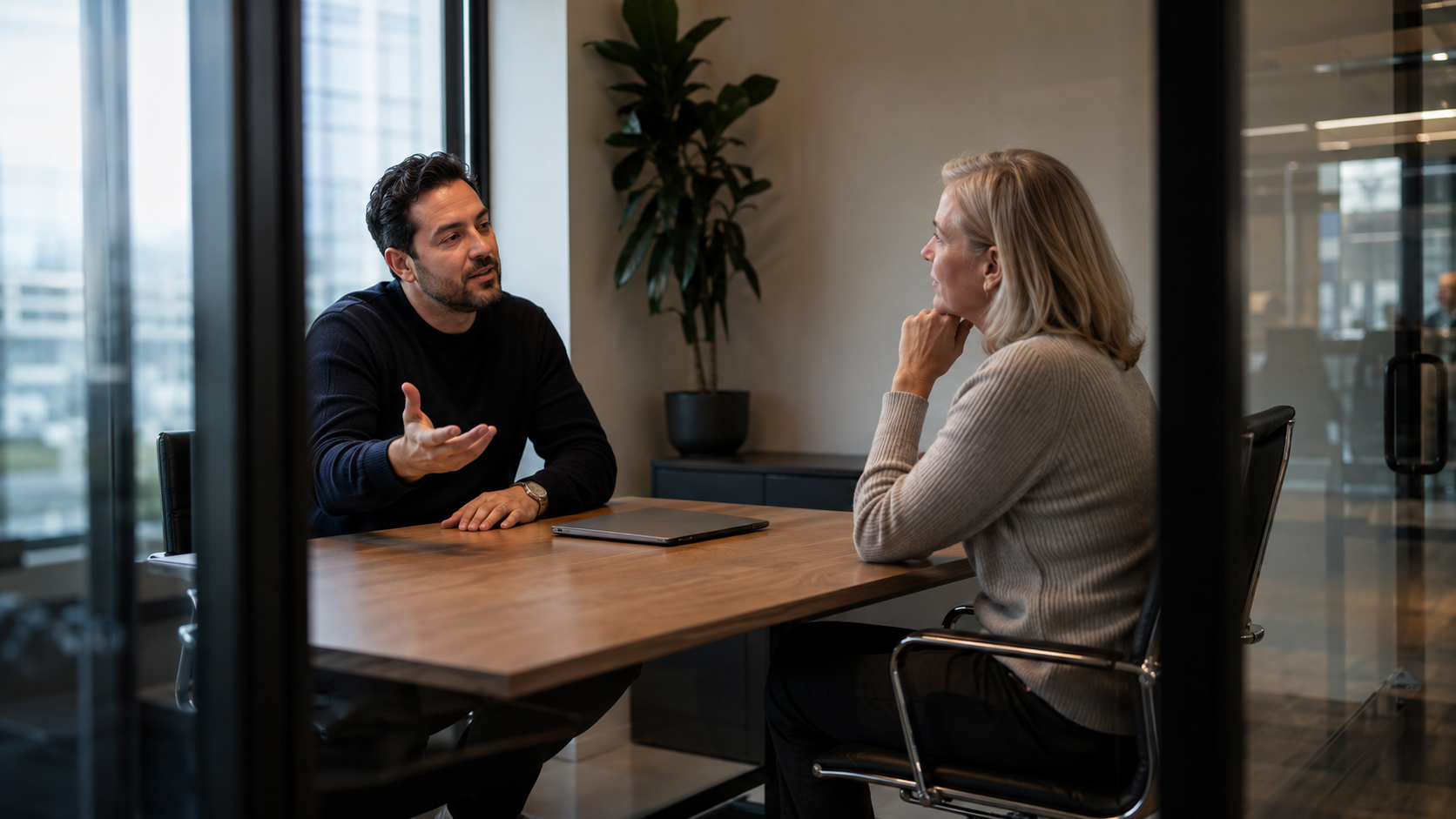 A sales manager and a seller seated across a small table in a glass-walled office, in an unhurried working conversation.